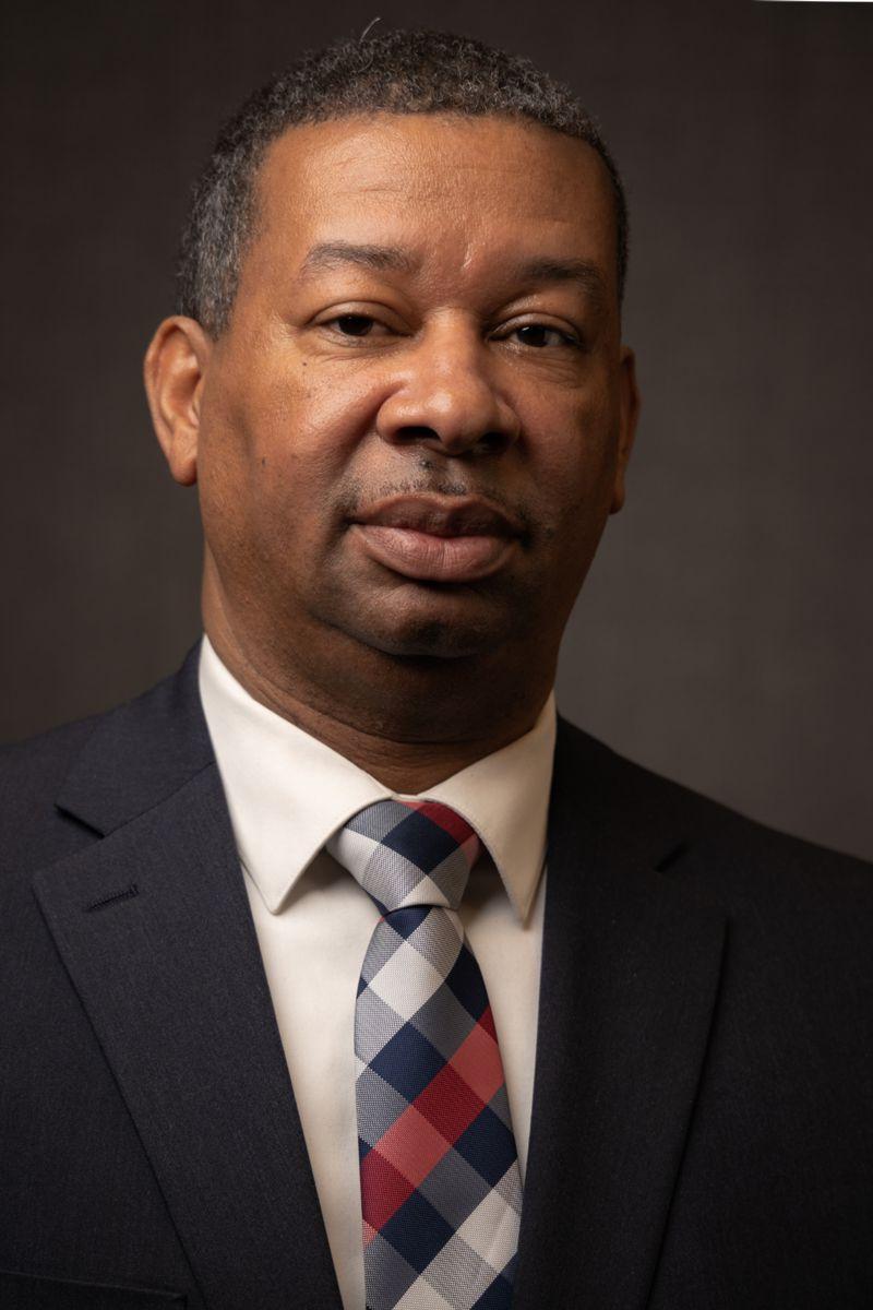Dr. 豪伊Gunby, 一名非裔美国男子，身穿灰色西装，内搭白领衬衫，系一条灰色和白色相间的红色条纹格子领带.